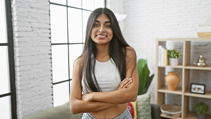 Smiling young indian woman with long brunette hair, standing in a modern living room, exuding...