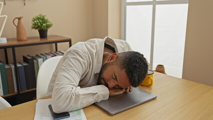 A tired young man with a beard is sleeping on his laptop in a modern apartment's living room