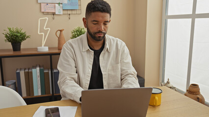 A focused african american man with a beard works on a laptop in a modernly decorated home office.