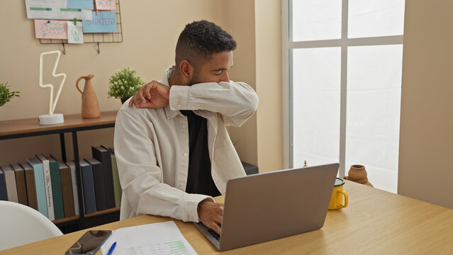 A young bearded african american man coughing into his elbow at his comfortable home workspace with laptop and coffee mug