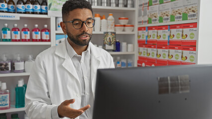 An attentive young black man wearing a lab coat in an indoor pharmacy setting, working on a computer.