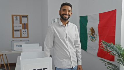 A smiling young man with a beard standing indoors near a mexican flag and voting booths,...