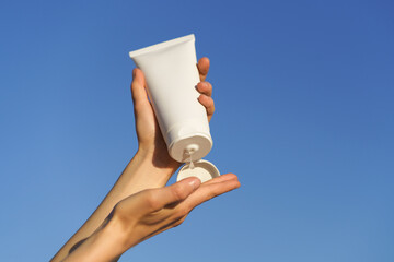 Female hands hold white mockup tube with cream or lotion and apply on palm against blue sky...