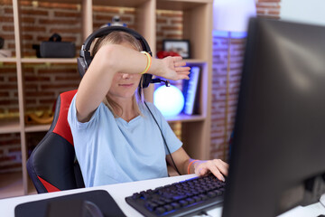 Young caucasian woman playing video games wearing headphones covering eyes with arm, looking...