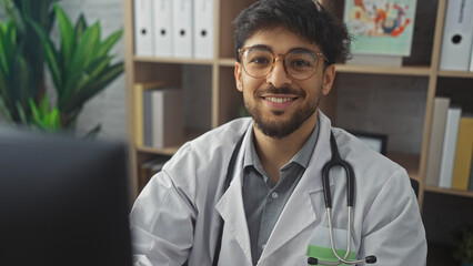 A smiling young arab man with a beard, wearing glasses and a lab coat, sits in a hospital office.