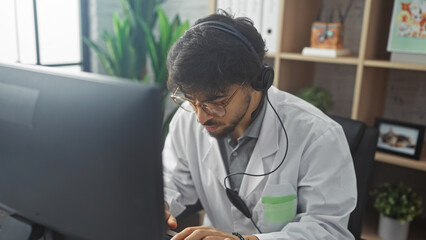 A focused young man with a beard working in a clinic office while wearing headphones and a lab coat.