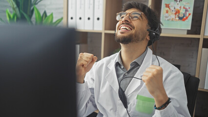 A cheerful man in a lab coat with headphones celebrates success in a bright office