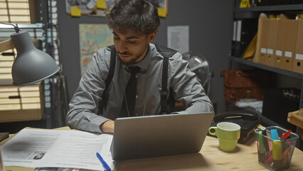 Handsome bearded man working at police station surrounded by documents, laptop, coffee mug