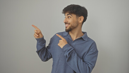 A young man with a beard in casual wear posing against a white background, smiling and pointing to...