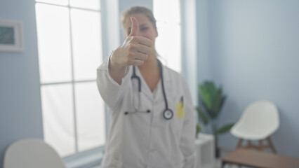 Fototapeta premium A positive young female doctor gesturing thumbs-up in a bright hospital room, symbolizing good health and recovery.
