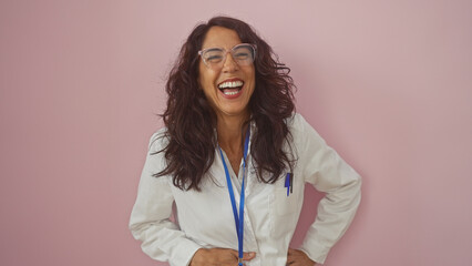 A joyful middle-aged woman in a white lab coat and clear glasses laughs heartily against a pink...