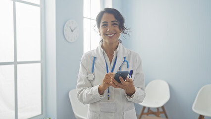 A smiling hispanic woman doctor holding a phone in a bright hospital room