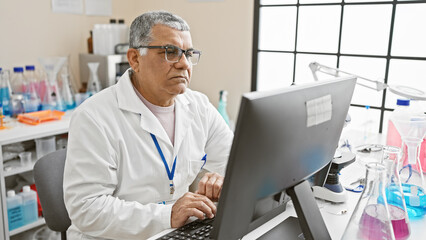 Mature grey-haired man works on computer in a modern laboratory setting, surrounded by scientific equipment.