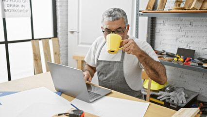 A mature man with grey hair sips from a cup while using a laptop in a well-equipped carpentry...