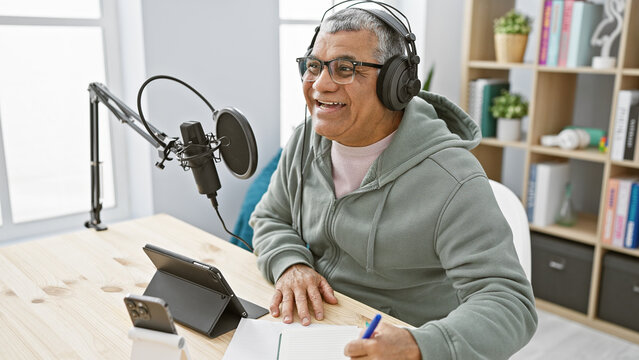 Smiling senior man with headphones speaking into a microphone in a modern radio studio setup.