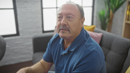 Middle-aged bald man with a moustache in a blue shirt sitting thoughtfully in a modern living room