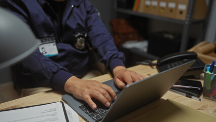 A young hispanic man working as a detective in a police station office, typing on a laptop at his...