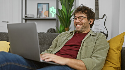 A cheerful young hispanic man with a beard wearing wireless earbuds using a laptop in a cozy living room.