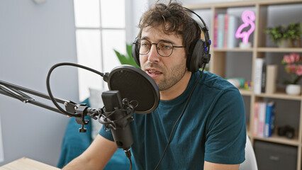 A young hispanic man with a beard speaks into a microphone in a radio studio, wearing headphones.