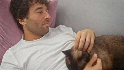 Relaxed young adult hispanic man lounging on a sofa indoors petting a siamese kitten