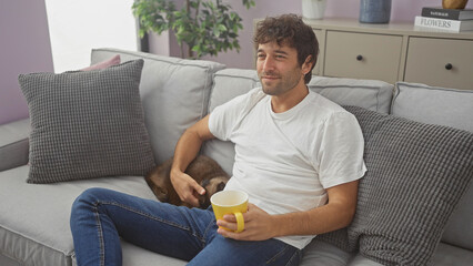 Relaxed young hispanic man petting a cat while sitting on a couch indoors with a mug in his hand.
