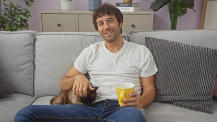 A smiling young hispanic man pets a cat while sitting on a living room couch with a cup in hand.