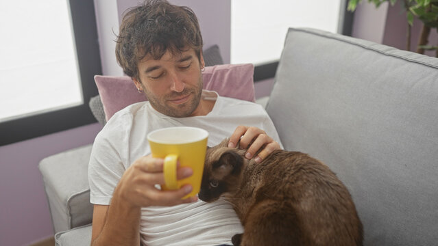 A relaxed young man enjoying a moment with his siamese cat while holding a coffee mug indoors.