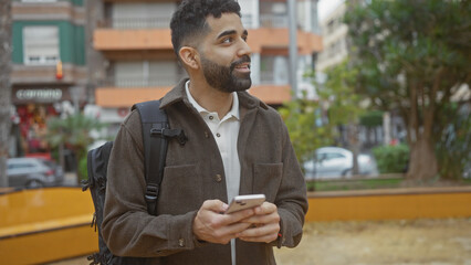 A handsome hispanic man uses a smartphone in a city park, portraying a casual urban lifestyle.