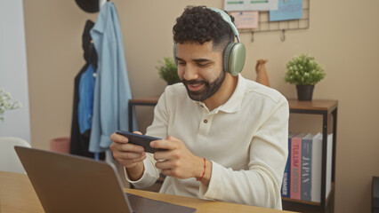 Hispanic man with beard using smartphone and laptop in living room, wearing headphones, smiling,...