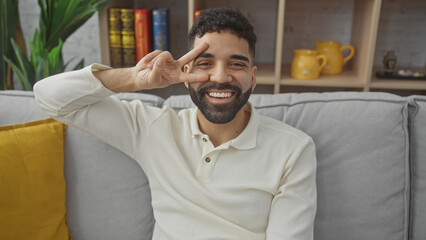 Handsome hispanic man making peace sign indoors, with a cozy living room backdrop.
