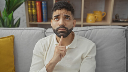 Pensive hispanic man with beard sitting on grey sofa in a cozy living room, contemplating.