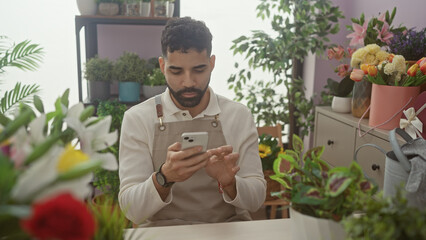 Hispanic man with beard using smartphone in a colorful flower shop interior filled with plants.