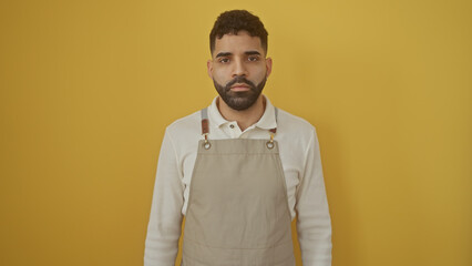 A handsome young hispanic man with a beard, wearing an apron, poses against an isolated yellow background.