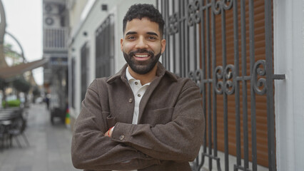 Smiling young hispanic man with beard standing arms crossed on urban city street
