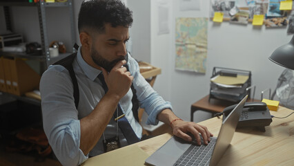 Hispanic detective working thoughtfully on laptop in cluttered office room