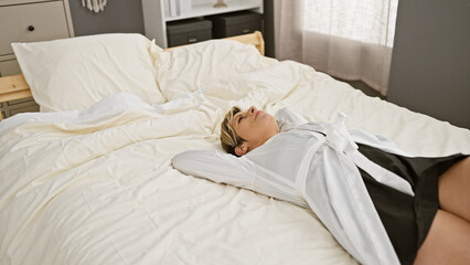 A young hispanic woman with short blonde hair relaxes on a bed inside a well-lit bedroom.