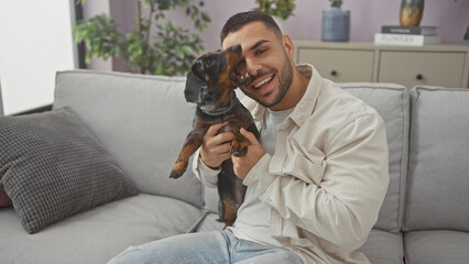 Smiling young hispanic man holding a dachshund dog in a cozy modern living room
