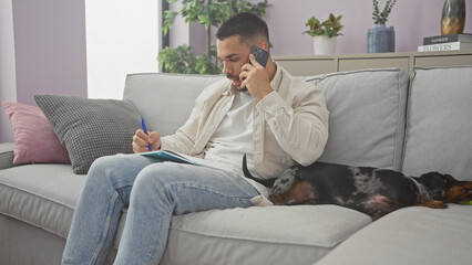 A young hispanic man multitasking with a phone call and notetaking, alongside a resting dachshund...