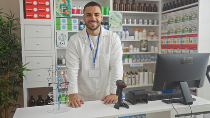 A smiling young hispanic man pharmacist stands confidently inside a well-stocked, modern pharmacy interior.