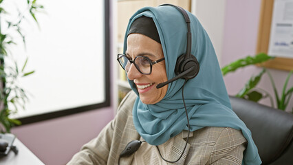 Smiling mature muslim woman with hijab using headset in modern office setting.