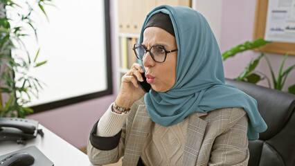 A professional woman in hijab speaks on a phone in a modern office setting, exuding concern and...