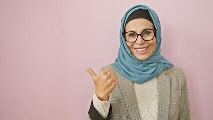 Smiling woman in hijab pointing sideways against a pink background, depicting positivity and...