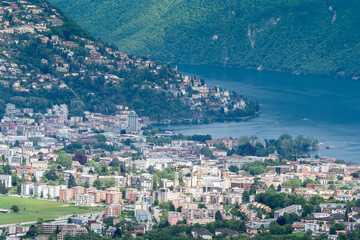 view of the town of Lugano in Italy. charming town, lush green hills, serene waters, lake lugano, colorful buildings, traditional italian architecture, waterfront, vibrant town square, bustling 