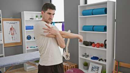 Hispanic man stretching at rehabilitation clinic against backdrop of exercise equipment and anatomy...
