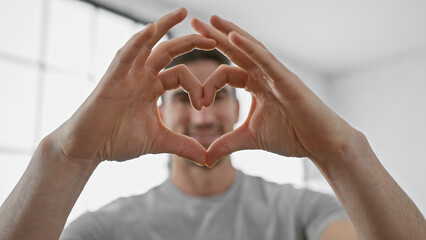 A young hispanic man forms a heart with his hands indoors, suggesting love, affection, and...
