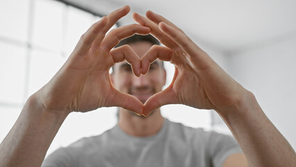 A young hispanic man forms a heart with his hands indoors, suggesting love, affection, and positivity.