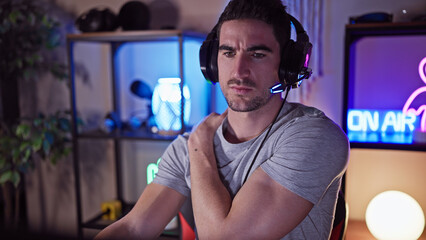 Handsome hispanic man with headphones in a dark gaming room looking pensive.