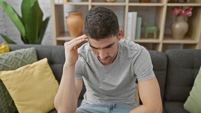 Thoughtful young hispanic man sitting indoors on a gray sofa in a cozy living room, contemplating