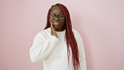 A pained african woman with braids touches her cheek against a pink background, signifying...