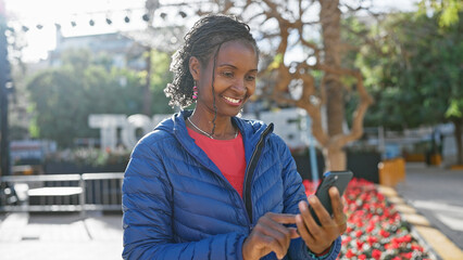 African woman smiling while using smartphone on sunny urban street.